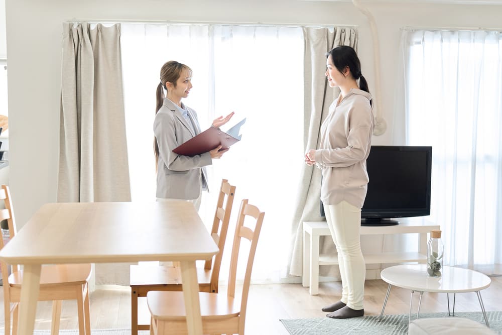 部屋の中で不動産担当者と話す女性。テーブルやテレビが置かれた明るいリビングルームの様子。